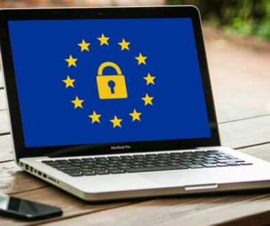 A laptop on a wooden table displays a blue screen with a yellow padlock surrounded by twelve yellow stars, symbolizing the European Union's GDPR. A smartphone and blurred outdoor background are also visible.
