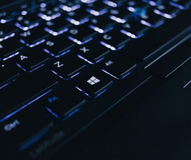 Close-up of an illuminated laptop keyboard in a dimly lit setting. The focus is on the Windows key, which is prominently lit among other keys like Shift, Z, X, and Alt. The lighting creates a blue-tinted glow across the keys, highlighting the keyboard's sleek design.