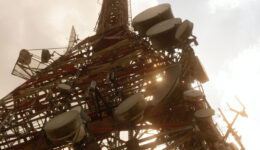 A tall communications tower is seen from below, silhouetted against the sky. Numerous large satellite dishes and antennas are attached to its metal framework. Sunlight peeks through the structure, casting a warm glow. Clouds are visible in the background.