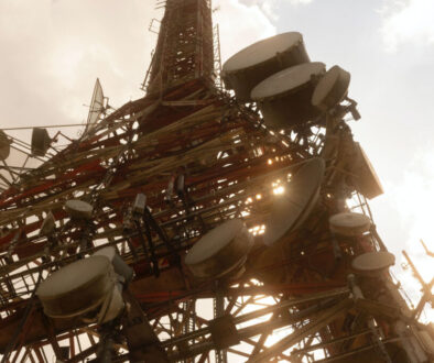 A tall communications tower is seen from below, silhouetted against the sky. Numerous large satellite dishes and antennas are attached to its metal framework. Sunlight peeks through the structure, casting a warm glow. Clouds are visible in the background.