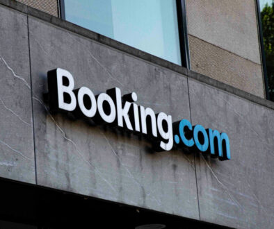 A gray building facade features the "Booking.com" logo, with "Booking" in white and ".com" in blue, both in a modern sans-serif font. The surface shows visible cracks and textures, hinting at the evolving landscape of digital markets. A window reflection is partially visible above the logo.