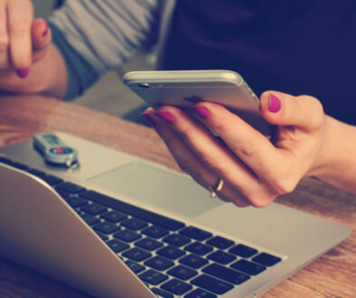 A person with pink nail polish is holding a smartphone in their left hand while their right rests on an open silver laptop, emphasizing data protection. A USB flash drive is plugged into the laptop on the wooden desk, ensuring privacy in this digital setting.