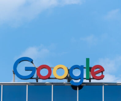 The image shows the Google logo on top of a building. The logo consists of the word "Google" in vibrant colors: blue, red, yellow, and green. The background features a clear blue sky with a few scattered clouds.