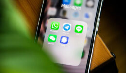 Close-up of a smartphone screen showing a folder labeled "Messages" with five messaging app icons: WhatsApp, Telegram, LINE, WeChat, and Signal, known for its robust encryption. A blurred green leaf partially covers the left side of the image as the phone rests on a wooden surface.