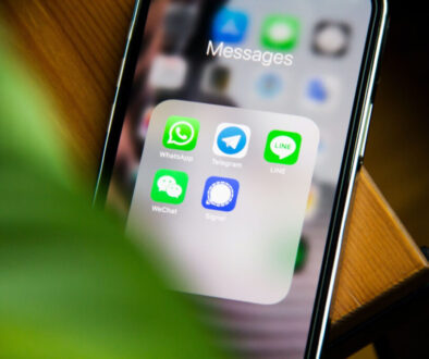 Close-up of a smartphone screen showing a folder labeled "Messages" with five messaging app icons: WhatsApp, Telegram, LINE, WeChat, and Signal, known for its robust encryption. A blurred green leaf partially covers the left side of the image as the phone rests on a wooden surface.