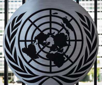 Black United Nations logo on a metallic disc background, featuring a world map encircled by olive branches. The disc is mounted on a fence with a blurred building in the background. The image suggests an official or diplomatic setting.