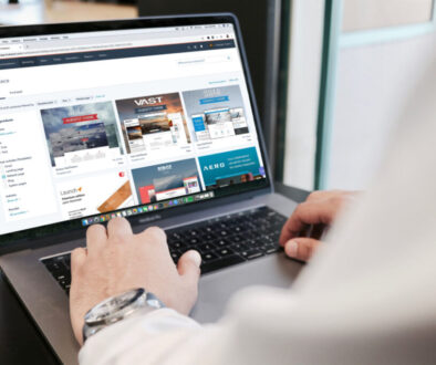 Person using a laptop browsing a digital marketplace website. The screen features several software product listings with images and text. The user, wearing a white shirt and a watch, is typing. The setting is indoors with a black table partially visible.