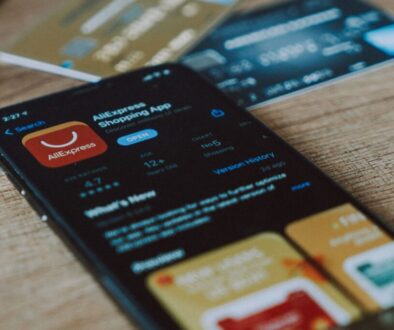 A smartphone displaying the Aliexpress Shopping App page in an app store, with the app's logo and Digital Services Act compliance rating visible. Resting on a wooden surface, blurred credit cards linger in the background. The screen shows app details including ratings and version history.