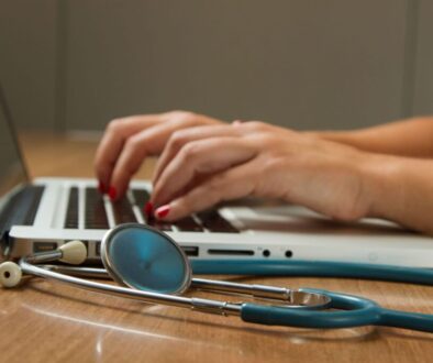 A person with red nail polish is typing on a laptop placed on a wooden surface. In the foreground, a teal stethoscope and documents about EHDS FAQs are resting on the table, suggesting a medical context linked to European Health Data Space initiatives. The background is blurred, focusing on the hands and stethoscope.