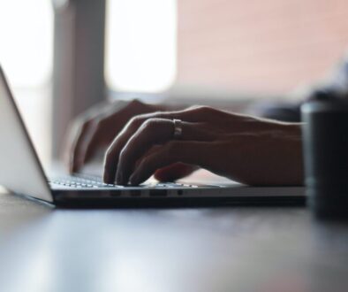 Close-up of hands typing on a laptop keyboard, with a ring adorning the right hand. The screen displays details about the AI Act approved by the European Council. The laptop rests on a wooden table, bathed in natural light from a window, creating a calm and focused atmosphere.