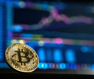A close-up of a physical gold Bitcoin coin is set against a blurred background displaying colorful financial charts on a computer screen. The coin, with its intricate patterns and logo, symbolizes cryptocurrency and digital finance trends amid evolving EU crypto regulation under MiCA.