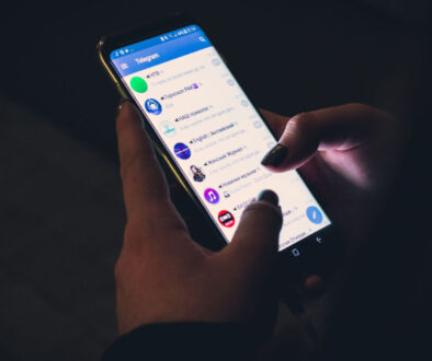 A person with dark nail polish is holding a smartphone, navigating the Telegram chat interface using their thumbs. The dimly lit screen shows a list of conversations with various contacts and groups, seamlessly integrating VLOP under the new Digital Services Act regulations.