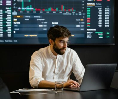 A man with a beard and glasses sits at a black table using a laptop, analyzing the impact of the AI Act. He's wearing a white shirt and is surrounded by documents. A glass of water is in front of him, while behind him, a large screen displays various stock market graphs and data in the dimly lit room.