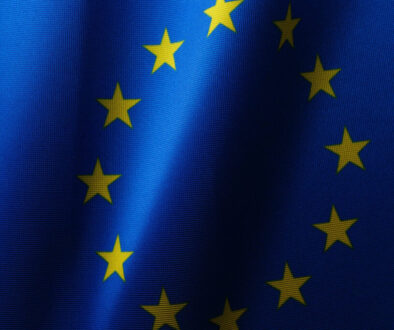 The image shows the flag of the European Union, featuring a circle of twelve gold stars on a blue background. The fabric appears to be slightly wavy, reflecting light and creating a sense of movement, much like the dynamic discussions led by the AI Act Monitoring Group.
