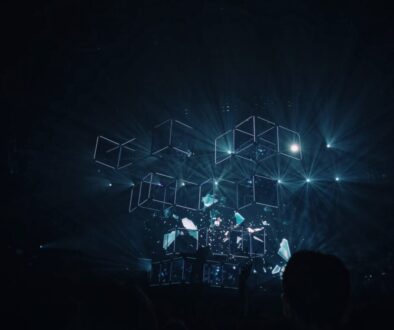 A dark stage is illuminated with blue light, showcasing a 3D geometric installation of illuminated cubes. Light beams radiate outward from the structure, creating a futuristic and immersive atmosphere. Silhouettes of people are visible at the bottom, indicating an audience.