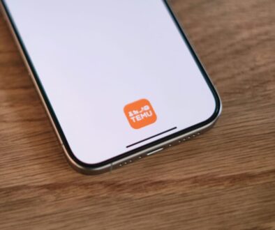 Smartphone on wooden surface displaying a white screen with an orange square icon labeled "TEMU" near the bottom. The phone's edges and charging port are visible, and the overall design suggests a modern device.