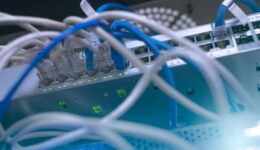 Close-up of network switches with multiple Ethernet cables plugged in. Green LED lights indicate active connections. The cables are mostly blue and white, and several ports on the metallic switch panel are occupied. The focus is on the cables and ports.