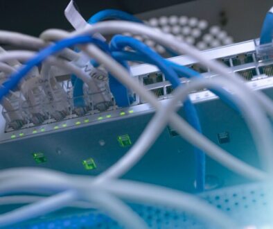 Close-up of network switches with multiple Ethernet cables plugged in. Green LED lights indicate active connections. The cables are mostly blue and white, and several ports on the metallic switch panel are occupied. The focus is on the cables and ports.