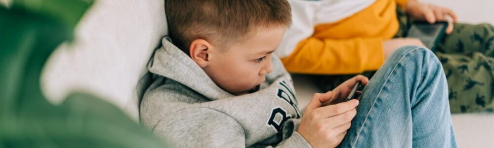 Two boys sit on a light-colored couch, both focused on handheld devices. The boy in the foreground wears a grey hoodie and blue jeans, holding a smartphone. The boy behind him wears a striped shirt with yellow and blue, looking at a tablet.