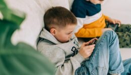 Two boys sit on a light-colored couch, both focused on handheld devices. The boy in the foreground wears a grey hoodie and blue jeans, holding a smartphone. The boy behind him wears a striped shirt with yellow and blue, looking at a tablet.