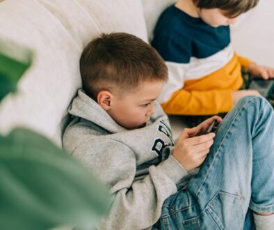 Two boys sit on a light-colored couch, both focused on handheld devices. The boy in the foreground wears a grey hoodie and blue jeans, holding a smartphone. The boy behind him wears a striped shirt with yellow and blue, looking at a tablet.