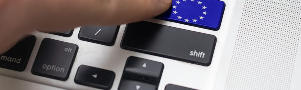 A close-up of a finger pressing a blue key with the European Union flag on a laptop keyboard. The blue key replaces the enter key and features a circle of yellow stars. Other keys, like shift and option, are also visible nearby.