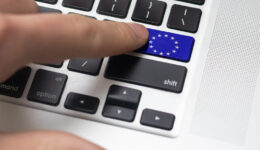 A close-up of a finger pressing a blue key with the European Union flag on a laptop keyboard. The blue key replaces the enter key and features a circle of yellow stars. Other keys, like shift and option, are also visible nearby.