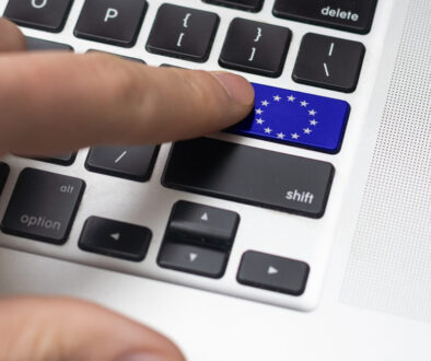 A close-up of a finger pressing a blue key with the European Union flag on a laptop keyboard. The blue key replaces the enter key and features a circle of yellow stars. Other keys, like shift and option, are also visible nearby.