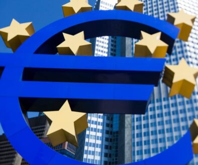 Large blue euro (€) symbol sculpture with yellow stars, representing the European Union, stands in front of modern glass skyscrapers under a clear blue sky. The buildings reflect light, emphasizing a financial or business district setting.