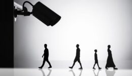 Silhouettes of four people, including a child, walk in a line from left to right against a light gray background. A large surveillance camera silhouette is positioned on the left, angled downwards toward the people, suggesting monitoring. The scene is reflected on a glossy surface below.