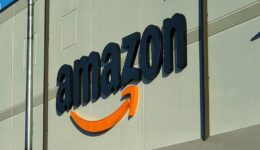 Large black “amazon” letters with an orange curved arrow beneath the text are mounted on a light gray building. The logo is partially illuminated by sunlight, with shadow and light dividing the wall. Vertical and horizontal lines accent the building’s exterior.