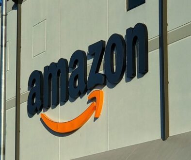 Large black “amazon” letters with an orange curved arrow beneath the text are mounted on a light gray building. The logo is partially illuminated by sunlight, with shadow and light dividing the wall. Vertical and horizontal lines accent the building’s exterior.