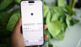 A hand holds a smartphone displaying the ChatGPT app with a white interface. The keyboard is open, and the screen shows options like “Design a database schema.” Large green leaves are blurred in the background.