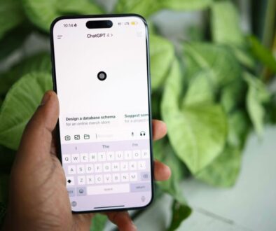 A hand holds a smartphone displaying the ChatGPT app with a white interface. The keyboard is open, and the screen shows options like “Design a database schema.” Large green leaves are blurred in the background.