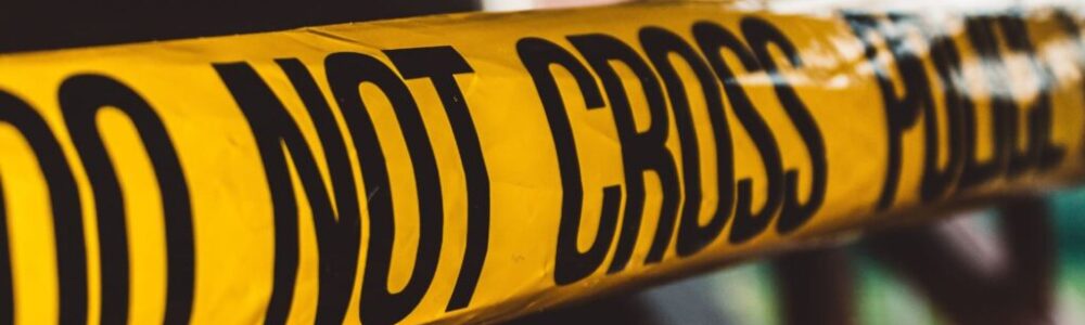 Close-up of several overlapping yellow police tape strips with bold black text reading "DO NOT CROSS." The tape is slightly wrinkled and creates a barrier, indicating a restricted or potentially hazardous area. The background is blurred and indistinct.