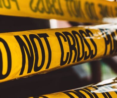 Close-up of several overlapping yellow police tape strips with bold black text reading "DO NOT CROSS." The tape is slightly wrinkled and creates a barrier, indicating a restricted or potentially hazardous area. The background is blurred and indistinct.