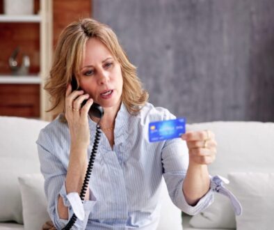 A woman with blonde hair sits on a white couch, holding a blue credit card in one hand while talking on a corded phone. She appears concerned or focused. A bookshelf and a gray wall are visible in the background.