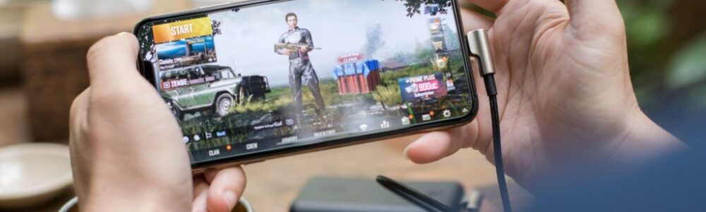 A person holds a smartphone horizontally, playing a shooting video game showing a character, vehicles, and supplies on the screen. The phone is plugged in with a charging cable. Background surfaces and objects are blurred, focusing on the hands and device.