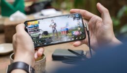 A person holds a smartphone horizontally, playing a shooting video game showing a character, vehicles, and supplies on the screen. The phone is plugged in with a charging cable. Background surfaces and objects are blurred, focusing on the hands and device.