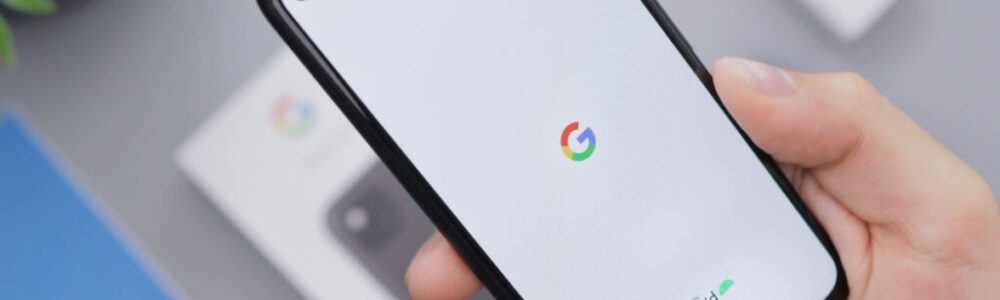 A hand holds a black smartphone displaying the Google logo and “powered by Android” on a white screen. A charger, phone box, and blue booklet are blurred in the background on a gray surface. A green plant is visible in the top left corner.