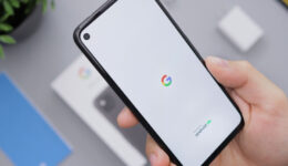A hand holds a black smartphone displaying the Google logo and “powered by Android” on a white screen. A charger, phone box, and blue booklet are blurred in the background on a gray surface. A green plant is visible in the top left corner.