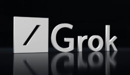 A 3D metallic logo with a slash in a square shape appears beside the word “Grok” in shiny silver letters. The logo and text are set against a glossy black background, with their reflections visible beneath them.