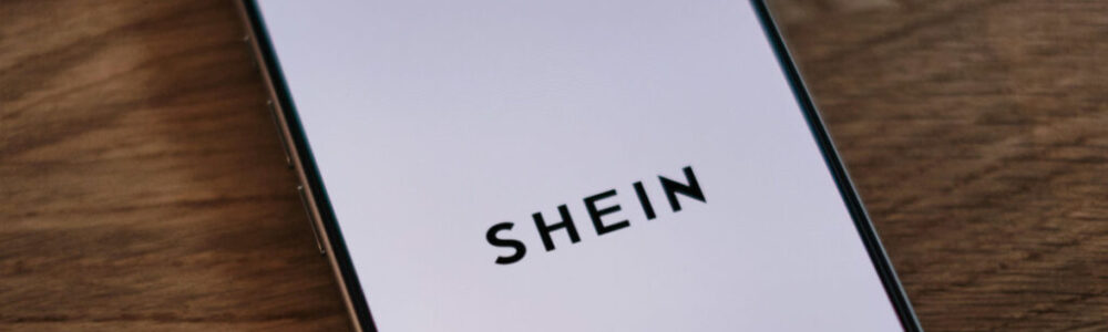 A smartphone displaying the SHEIN logo on its screen is placed diagonally on a wooden surface. The phone screen is white with the black text "SHEIN" centered on it. The wooden surface shows a natural grain texture.