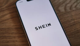 A smartphone displaying the SHEIN logo on its screen is placed diagonally on a wooden surface. The phone screen is white with the black text "SHEIN" centered on it. The wooden surface shows a natural grain texture.