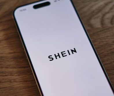 A smartphone displaying the SHEIN logo on its screen is placed diagonally on a wooden surface. The phone screen is white with the black text "SHEIN" centered on it. The wooden surface shows a natural grain texture.