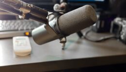 A professional broadcast microphone on an adjustable arm is positioned in the foreground. In the soft-focus background, a computer monitor, a keyboard, and a white remote control sit on a desk.