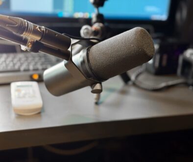A professional broadcast microphone on an adjustable arm is positioned in the foreground. In the soft-focus background, a computer monitor, a keyboard, and a white remote control sit on a desk.