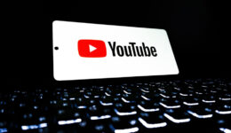 youtube A smartphone displaying the YouTube logo and name in bold letters floats above a glowing, out-of-focus computer keyboard with illuminated keys in a dark environment. The scene suggests digital media, streaming, or online video content creation.