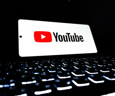 A smartphone displaying the YouTube logo and name in bold letters floats above a glowing, out-of-focus computer keyboard with illuminated keys in a dark environment. The scene suggests digital media, streaming, or online video content creation.