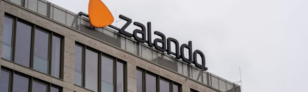 A modern building with large windows features a sign for Zalando, including its logo—an orange, rounded triangle—mounted on the rooftop against an overcast sky. The sign is in bold, black letters.